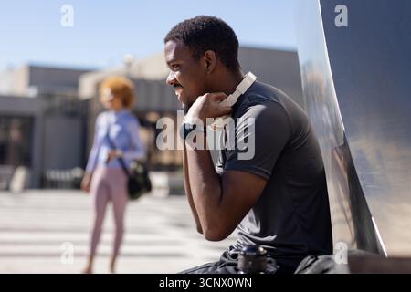 African american man adjusting headphones, checking smartwatch by panel in plaza, copy space Stock Photo