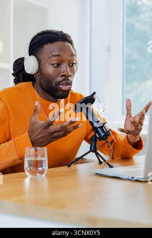African American man wearing orange shirt and headphones speaking into microphone at home office Stock Photo