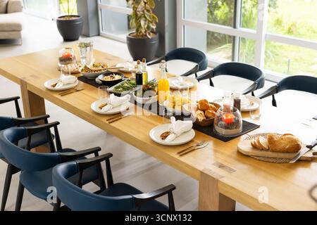 Wooden dining table hosting place settings, salads and breads, juice bottle, candles, navy chairs Stock Photo
