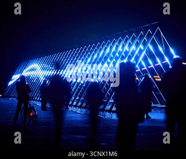 Blue led triangles form a striking geometric tunnel at night, with silhouetted figures walking through. The fusion of technology and human presence Stock Photo