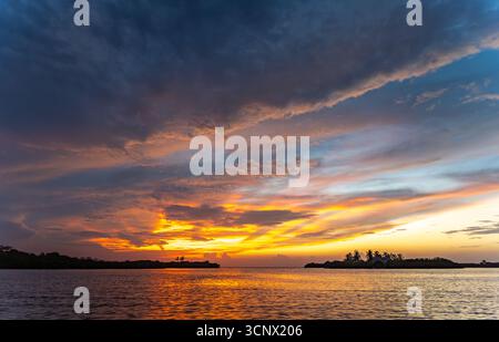 Stunning sunset over the Caribbean Sea with dramatic clouds and golden light. Small islands with palm trees appear on the horizon, adding to the tranq Stock Photo