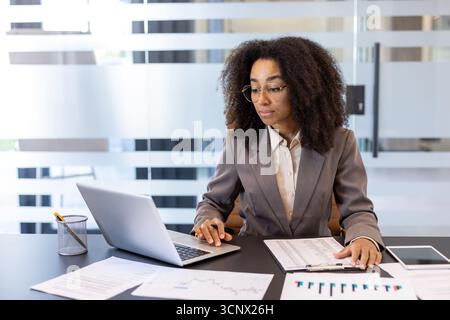 Young african american female accountant, financial expert sitting in office at desk and working on laptop and documents. Stock Photo