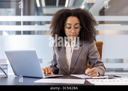 Serious and concentrated young African-American woman sitting in the office at the table with a laptop, working with documents, filling in data and tables. Stock Photo