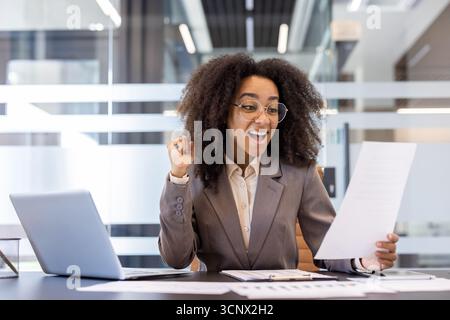 A happy young African-American business woman is sitting in the office at the desk with a laptop and is happy looking at the documents ,she is holding in her hand. Stock Photo