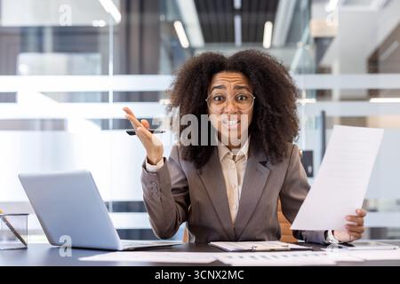 Portrait of angry and disappointed African American woman sitting in office at workplace, holding papers and gesturing emotionally to camera. Stock Photo