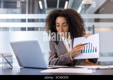 A young African-American businesswoman sits in the office at the table in front of a laptop and conducts an online conference, shows work charts to the camera. Stock Photo