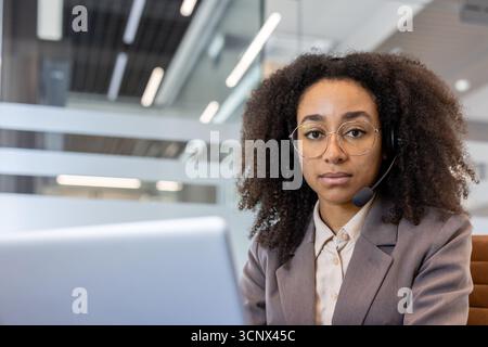 Close-up portrait of a young African-American woman in a headset working on a laptop, sitting at a table and looking seriously into the camera. Stock Photo