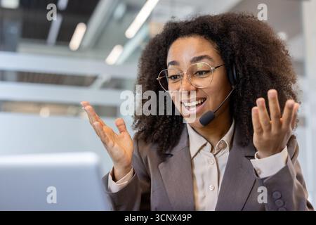 A young smiling afro-american woman conducts an online meeting in the office using a laptop, sitting at a desk in a headset and suit, gesturing with her hands to the camera. Stock Photo