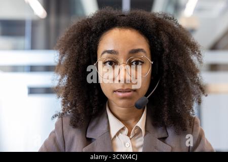 Close-up portrait of young serious and concentrated African-American woman in headset standing in office and looking at camera. Stock Photo