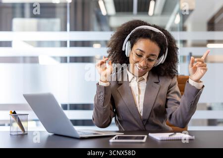 Young happy and relaxed African American woman sitting at desk in office wearing headphones, listening to music, dancing while sitting in chair. Stock Photo