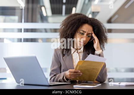 Upset young African American woman sitting at the desk in the office, holding her head with her hand and reading received letter with news and documents. Stock Photo