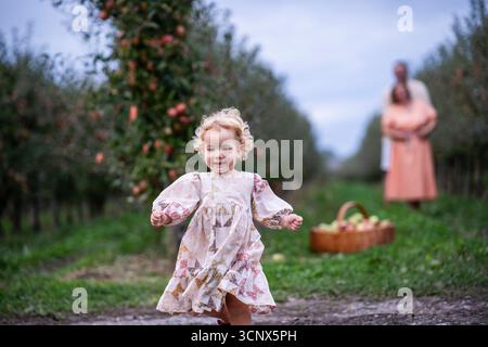 A joyful child runs through an apple orchard, while a couple stands in the background. A basket brimming with freshly picked apples completes the whol Stock Photo