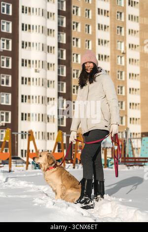 A woman in a warm coat and knit hat enjoys a snowy winter day with her Labrador retriever. The duo stands happily in a snow-covered park, surrounded b Stock Photo