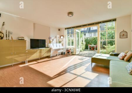 A spacious modern living room featuring sunlight streaming through large windows, lush greenery visible outside, and cozy furnishings creating a warm Stock Photo