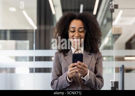 Close-up photo of a young African-American curly-haired smiling business woman inside an office and using a mobile phone. Stock Photo