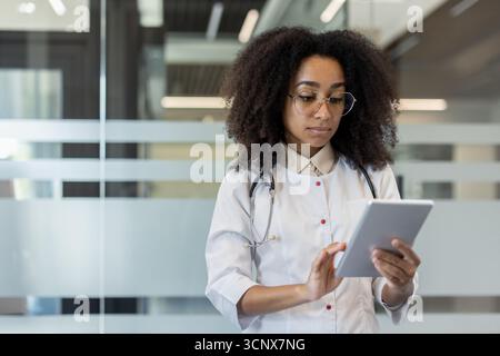 A young African-American female doctor in a white coat stands in the lobby of the clinic and concentrates on using a tablet. Stock Photo