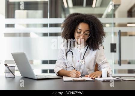 Young smiling African American female doctor and nurse sitting in clinic at desk with laptop, taking notes, working on laptop. Stock Photo