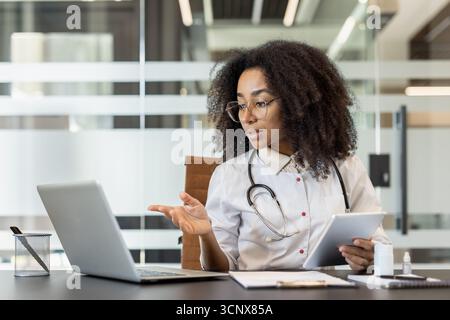 Concentrated and serious young afro-american female doctor sitting in clinic office at table in front of laptop, holding tablet in hand and consulting patient online, conducting video conference. Stock Photo