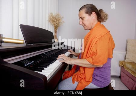 A middle-aged woman wearing an orange scarf plays the piano at home. The setting conveys a sense of tranquility and enjoyment as she engages in her mu Stock Photo