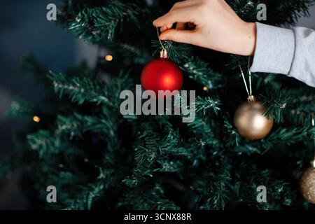 A hand carefully places a red ornament on a beautifully decorated Christmas tree. The green branches and other golden ornaments create a festive and j Stock Photo
