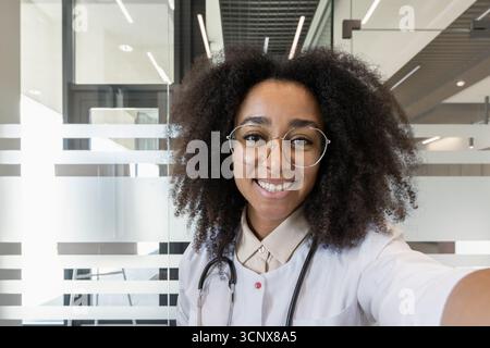 Healthcare professional smiling while taking selfie in office. Person wearing glasses and stethoscope, conveying professionalism and friendliness. Capturing moment of connection using smartphone. Stock Photo