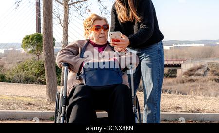 A warm scene of a grandmother in a wheelchair and her granddaughter enjoying time together outdoors. The granddaughter shows something interesting on Stock Photo