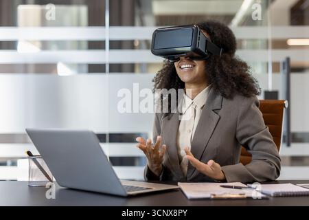 Businesswoman in office using virtual reality headset while interacting with laptop. Engaged in immersive technology experience, showcasing innovation, creativity, and modern workplace advancements. Stock Photo