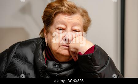 A close-up of an elderly woman resting her head on her hand, wearing a warm jacket indoors. Her facial expression conveys introspection, fatigue, or q Stock Photo