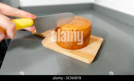 Close-up of a person using a knife with a yellow handle to cut a round cheese wheel on a wooden board. The countertop is gray, providing a clean kitch Stock Photo