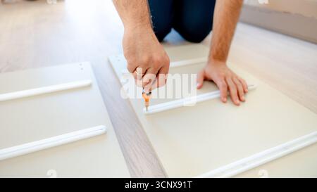 Cropped unrecognizable man with a wedding ring uses a screwdriver to assemble furniture. The focus is on hands working meticulously on light-colored w Stock Photo