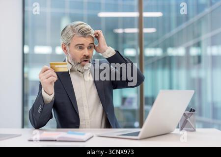 Stressed mature businessman looking at laptop, holding a credit card, and having a shocked expression on his face while experiencing online shopping payment issues Stock Photo