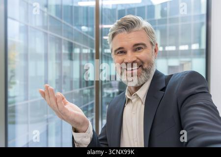 Mature businessman with a gray beard and professional attire, standing in a brightly lit modern office building with glass windows, smiling and gesturing with an open hand Stock Photo