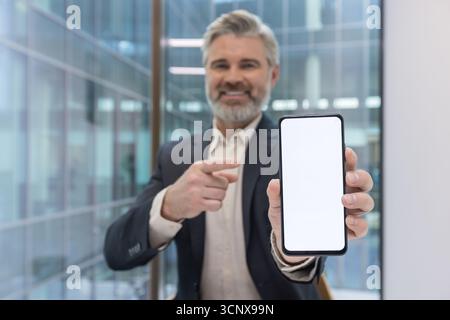 Senior businessman in a suit smiling and pointing at a smartphone with a blank white screen in a modern office, presenting a mobile app mockup for business promotion Stock Photo