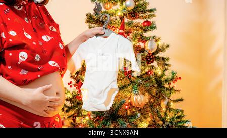 Cropped unrecognizable mother, faceless and anonymous, shares holiday joy by holding a onesie next to a decorated Christmas tree, celebrating pregnanc Stock Photo