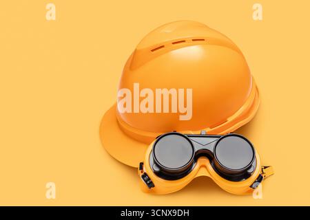 Close-up image of an orange hard hat and protective goggles set against a matching orange background. Represents safety equipment used in construction Stock Photo