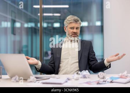 Mature businessman shrugging, feeling confused and frustrated, sitting at his messy office desk amidst crumpled papers, having writer's block and experiencing work-related stress Stock Photo
