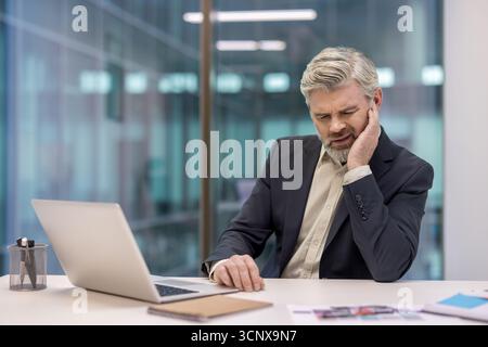 Senior businessman experiencing discomfort and pain from a toothache, holding his cheek while focusing on a laptop during working hours in a modern office environment Stock Photo