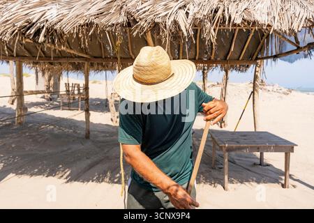 A man wearing a straw hat sweeping sand at Punta Astilleros Beach, Colombia. The rustic setting with wooden structures highlights a serene coastal env Stock Photo