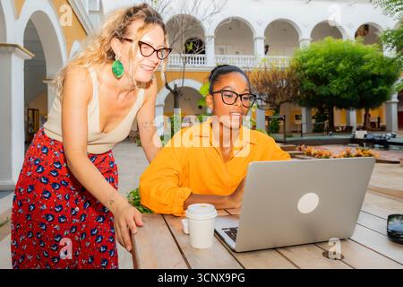 Multiethnic young women working as digital nomads. A caucasian woman smiles while a black woman focuses on her laptop. They enjoy a relaxed and produc Stock Photo