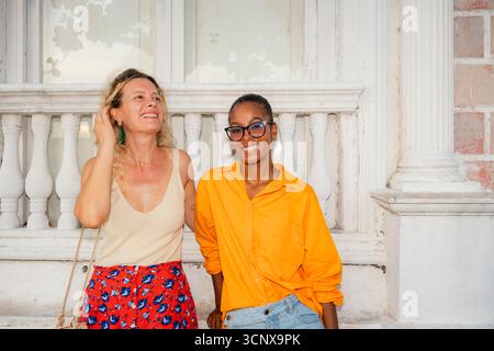 Young multiethnic women, a Caucasian and a Black woman, smiling joyfully outside a historic building with classic architecture, enjoying a sunny day i Stock Photo