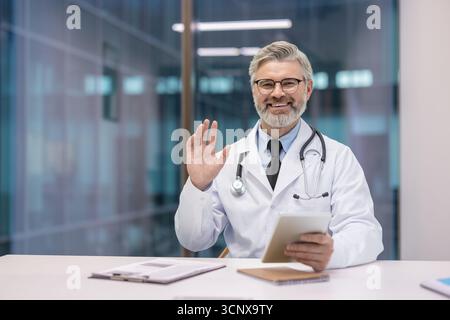 Older smiling doctor in white coat and stethoscope waving hello during a virtual medical appointment, offering care and advice through a digital tablet Stock Photo