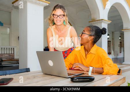 Two multiethnic women collaborate as digital nomads at an outdoor workspace. One woman stands and smiles while the other sits, working on a laptop, bo Stock Photo