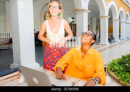Two young multiethnic women, a caucasian and a black woman, enjoy working and relaxing together outdoors, smiling and laughing with a laptop on the ta Stock Photo