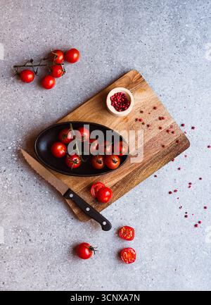 Fresh cherry tomatoes are arranged on a cutting board next to a serrated knife, ready to be sliced for a vibrant tomato salad. A bowl of peppercorns a Stock Photo
