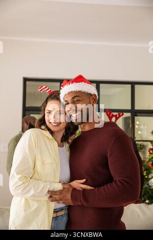 Diverse couple smiling at home in santa hat, candy-cane antler headband by christmas tree Stock Photo