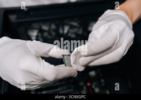 Gloved hands of male technician cleaning computer processor with white cloth, preparing for installation in computer case, ensuring optimal Stock Photo