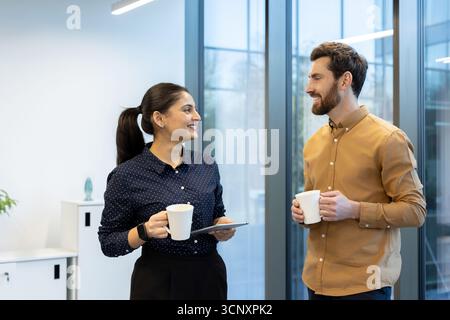 Two diverse business colleagues engaging in a friendly discussion and collaborating, holding coffee mugs during a casual break in a modern office environment Stock Photo