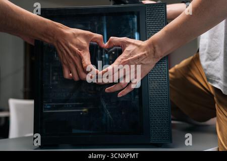 Two technicians making heart shape with hands in front of computer case, expressing passion and care for technology and computer maintenance, close-up Stock Photo