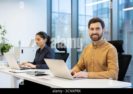 Smiling businessman working on laptop at his desk in a modern office, while a female colleague works in the background, representing collaboration and professional productivity Stock Photo