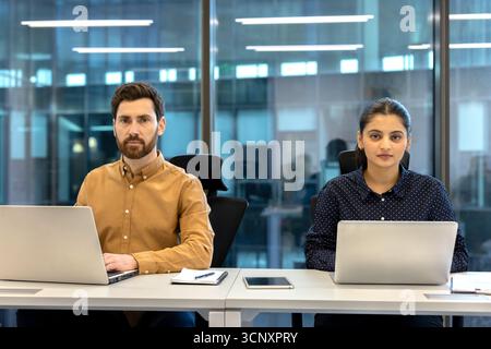 Two focused business professionals, a man and a woman, are working side-by-side on laptops at their shared desk in a modern corporate office environment, looking directly at the camera Stock Photo